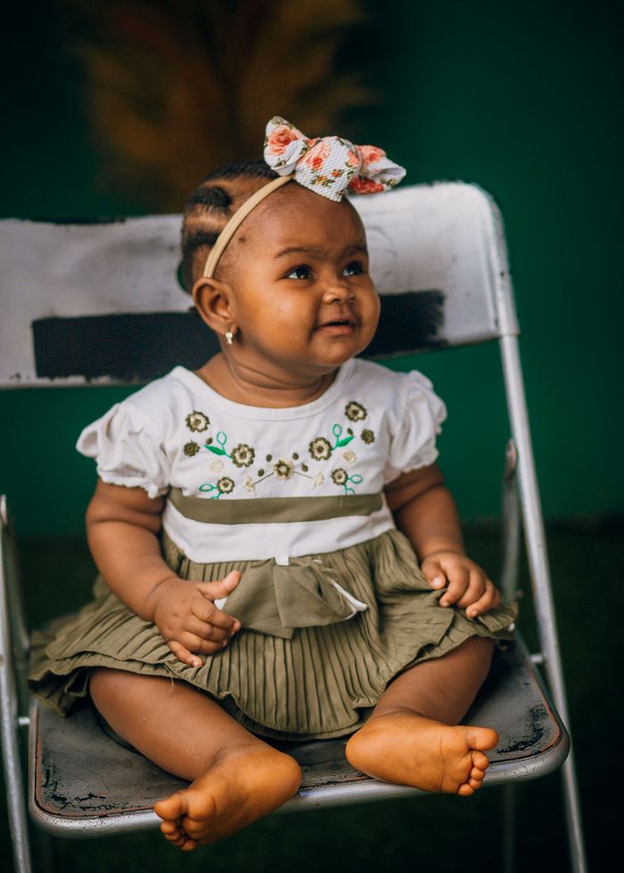 Charming baby girl with headband, sitting on chair, smiling warmly.